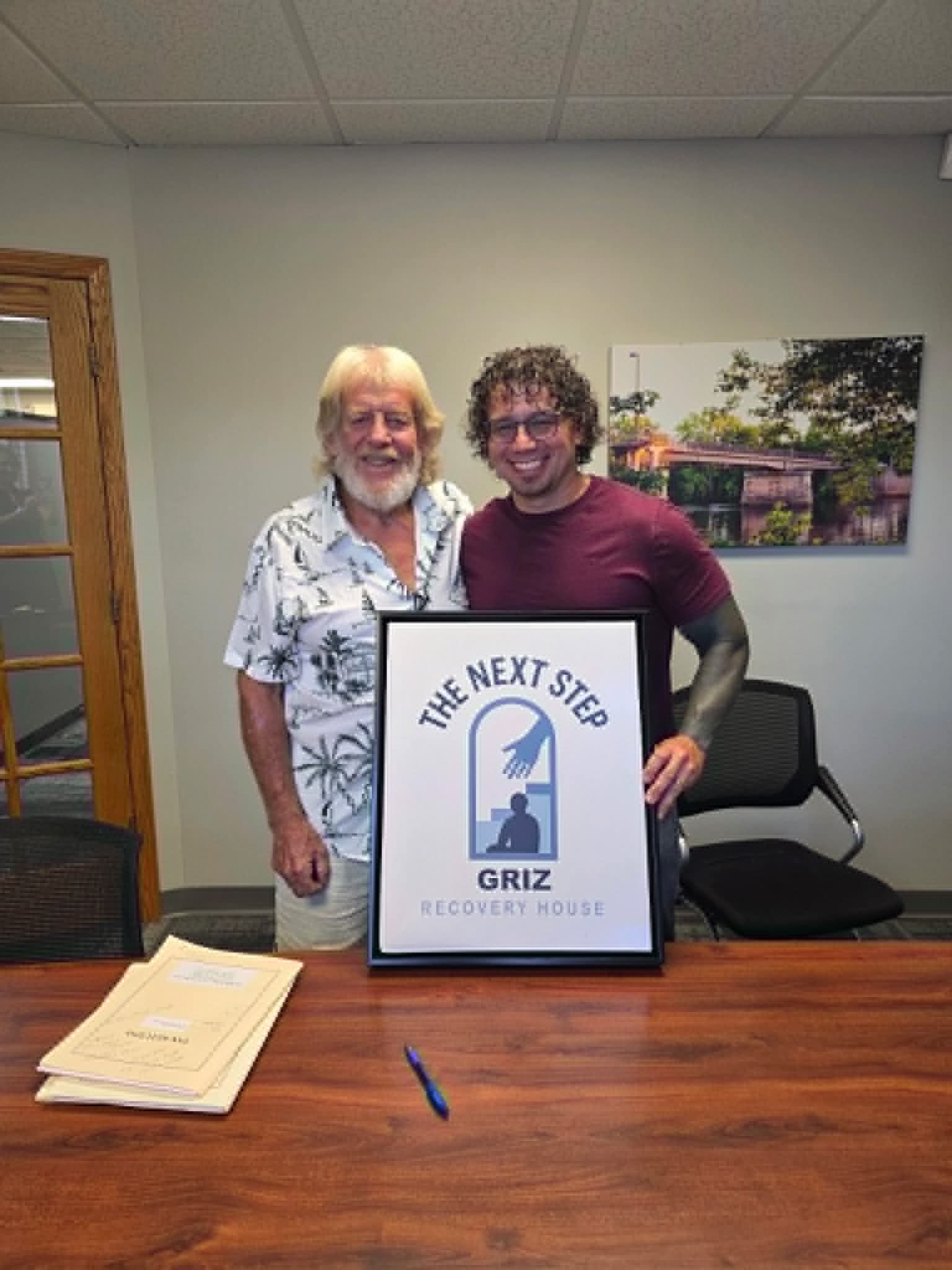 Two people standing behind a framed Griz Recovery House sign